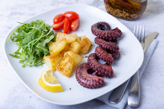 Grilled octopus with fried potatoes, arugula, tomatoes and lemon. Close-up.