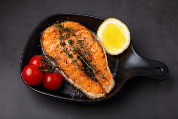 Grilled salmon with cherry tomatoes thyme and lemon on a small black grill pan Fried fish is a traditional dish of home cooking and restaurants Black background selective focus closeup