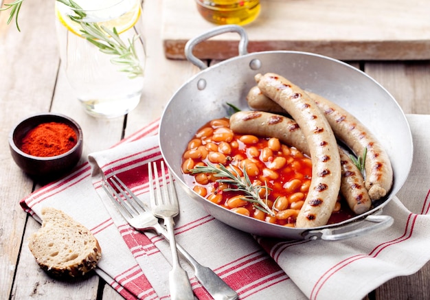 Grilled sausages with beans in tomato sauce in with rosemary and paprika in a wintage pan on a wooden background