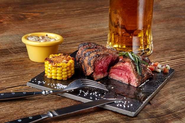 Grilled steak with corn with mushrooms sauce on cutting board and mug of beer on wooden table