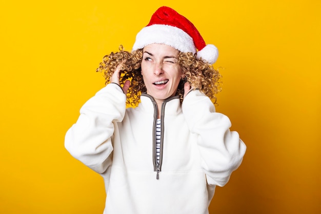 Grimacing young woman in a santa claus hat straightening curly hair on a yellow background.