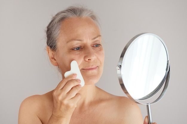 Grizzled mature woman massaging her face with a jade board while looking in the mirror