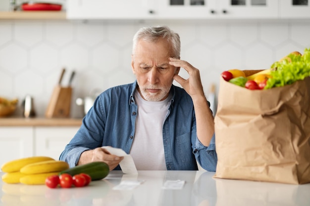 Grocery expenses upset senior man in kitchen checking bill after food shopping