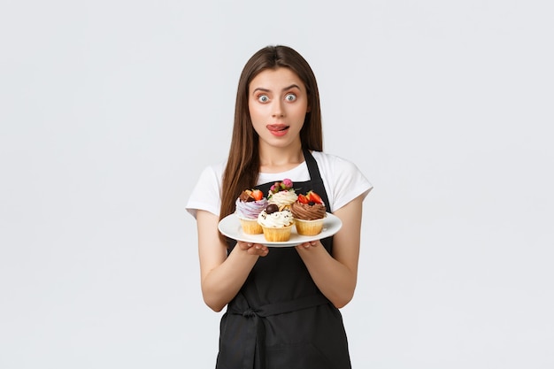 Grocery store employees, small business and coffee shops concept. Cute young female barista in black apron holding tasty dessert, lick lips desire have bite of delicious cupcakes, white background.