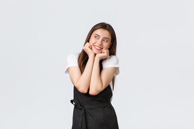 Grocery store employees, small business and coffee shops concept. Dreamy adorable barista imaging or thinking about something silly, looking left and smiling cute, wear black apron