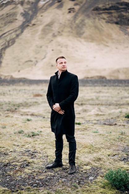 Groom in a black coat on a background of a mountain. Destination Iceland wedding.