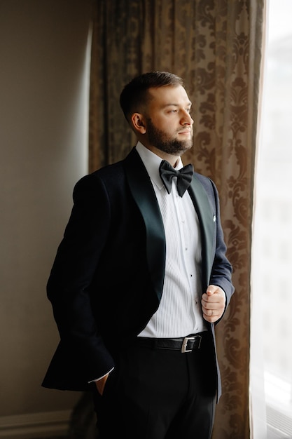 A groom in a black tuxedo stands in front of a window.