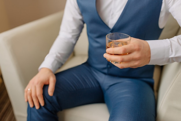 Groom, businessman sits and holds a glass of whiskey. Wedding morning groom preparation. Beautiful smiling man, groom posing and preparing for wedding. Close-up