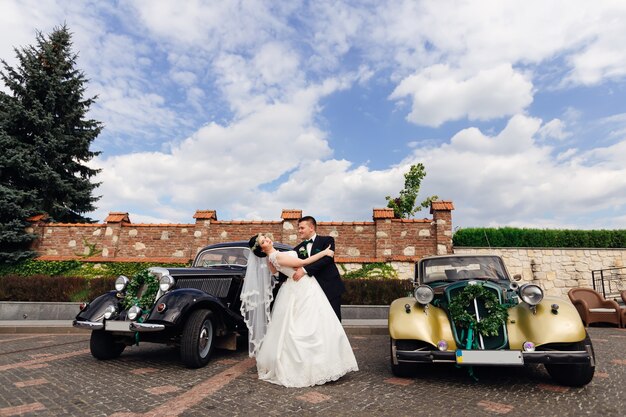 Photo the groom embraces the bride and she bends back two beautiful retro cars decorated with a wreath