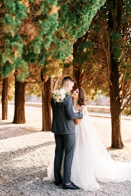 Groom hugs bride by the shoulders looking into her eyes while standing in a green cypress alley side