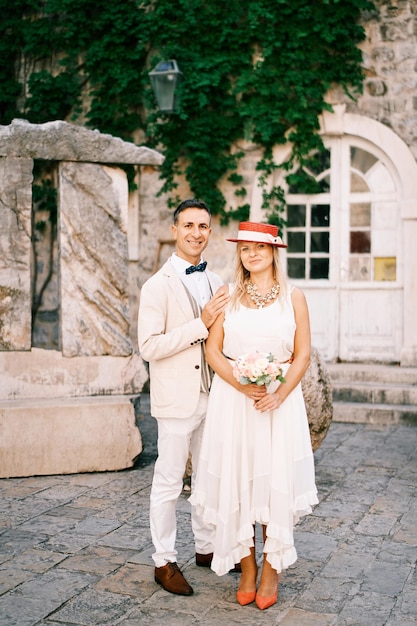 Groom hugs bride by the shoulders while standing on the paving stones near an old house