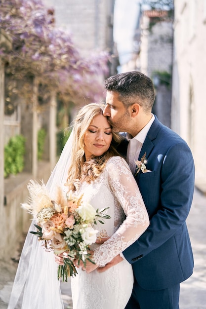 Groom hugs bride from behind and kisses her on the forehead near the garden fence