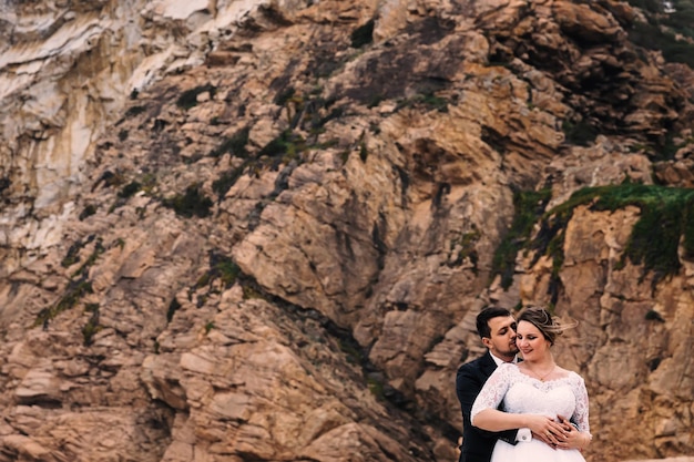 the groom hugs the bride from the side of the back and they close their eyes against the backdrop of the stone cliff close up