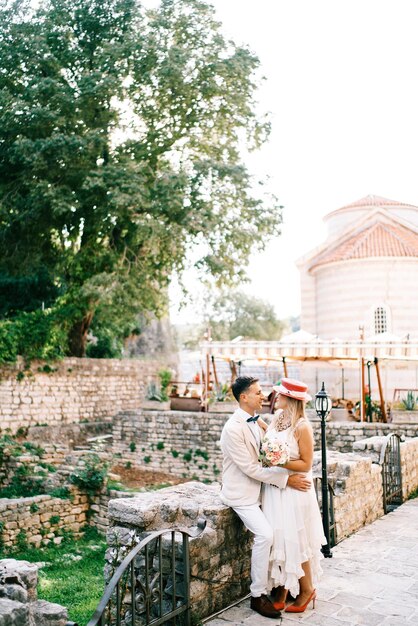 Photo groom hugs bride while sitting on a stone fence in the garden