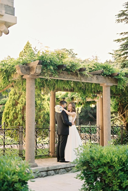 Groom hugs bride with a bouquet around the waist standing in a pergola in the garden