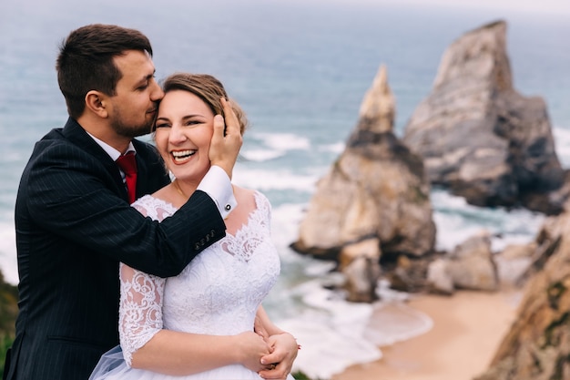 The groom kisses the bride and she sincerely smiles on  the backdrop of the ocean and cliffs.