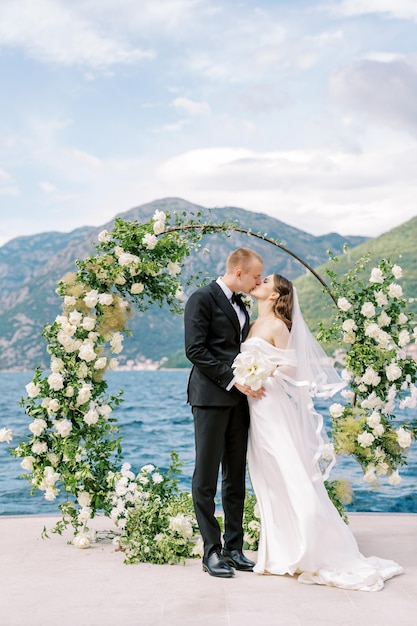 Groom kisses bride with bouquet at wedding arch on pier