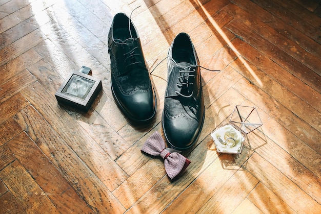 Groom's shoes standing on the wooden floor Perfumes with bowtie are nearby Wedding rings Stylish vintage photo