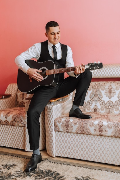 The groom sings songs to the guitar in the morning in his room Groom in a black suit Happy man Preparation for the wedding