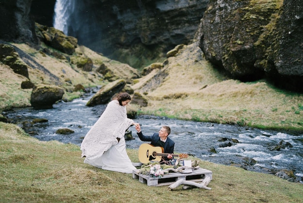 Groom sits with a guitar next to bride standing in a blanket on the river bank