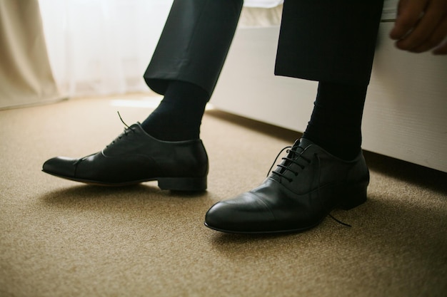 The groom sitting on the couch and tied shoelaces on black leather shoes, closeup shot. wedding