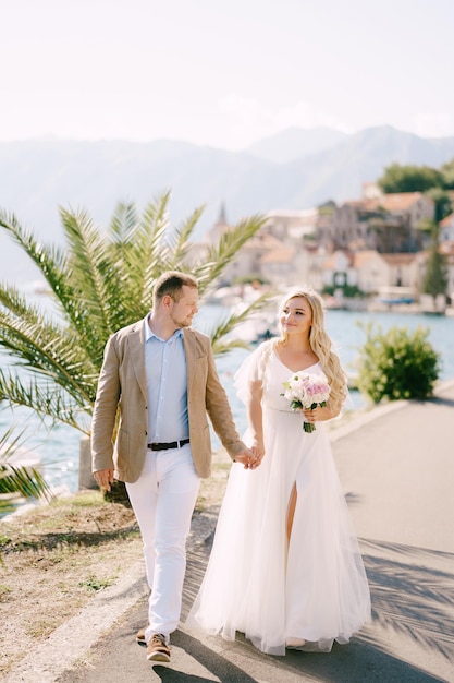 The groom and the smiling bride with a bouquet walk holding hands on the pier near the palm tree