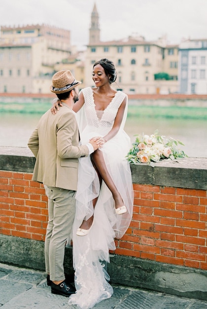 Groom stands near bride sitting on a brick fence on the river bank