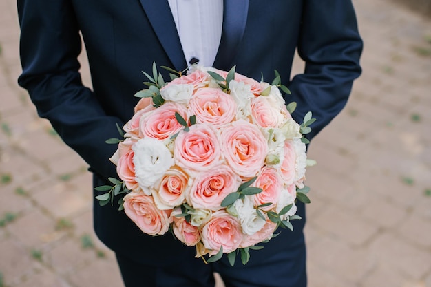 The groom stands with a wedding bouquet of roses and holds a jacket