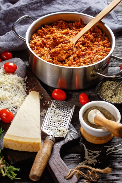 Ground beef risotto in a metal stewpot with a spoon on a dark wooden table with ingredients, italian cuisine, vertical view from above