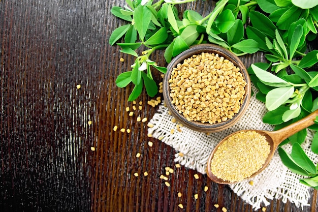 Ground fenugreek in a spoon and whole in a bowl on burlap with green leaves against dark wooden board on top