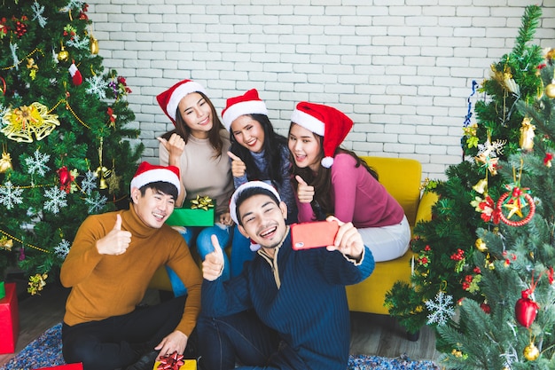 Group of asian friends taking selfie with friend together by smartphone at home during chrismas eve party or New year celebrate party. Happy winter Xmas and Happy new year party concept