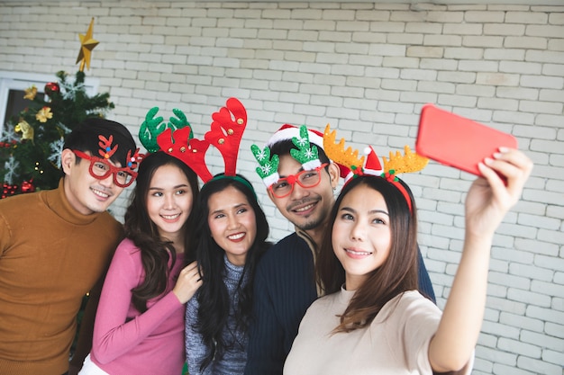 Group of asian friends taking selfie with friend together by smartphone at home during chrismas eve party or New year celebrate party. Happy winter Xmas and Happy new year party concept