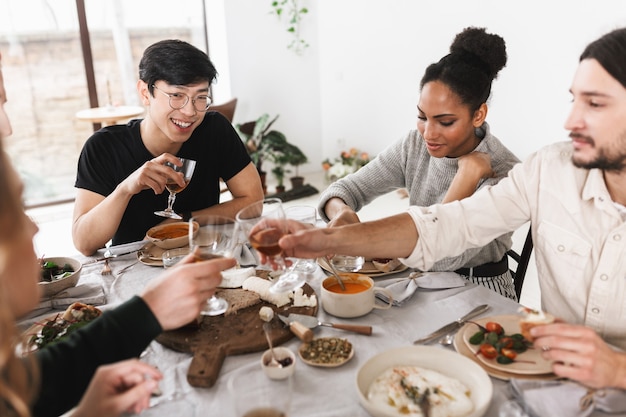 Group of attractive international friends sitting at the table full of food happily spending time together