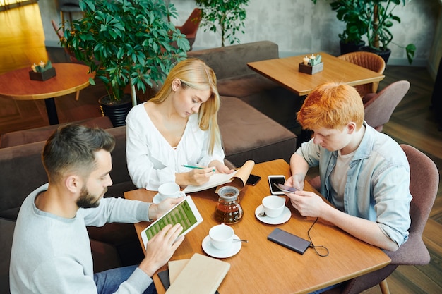 Group of busy students sitting by table in cafe after classes, guys using gadgets while blonde girl making notes in notepad