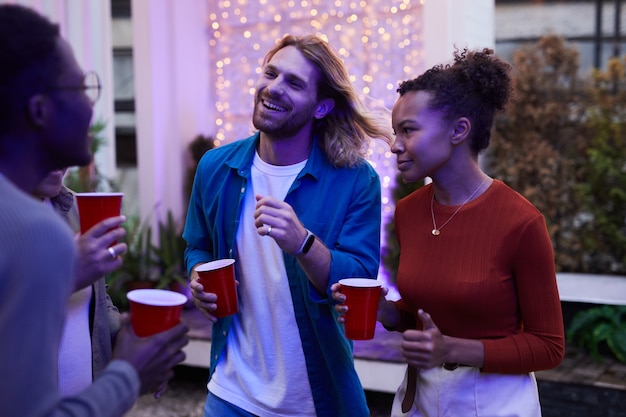 Group of carefree young people enjoying night party outdoors at rooftop lit by colored lights