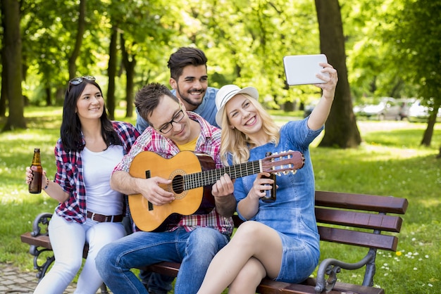 Group of caucasian friends playing guitar and taking selfies on a park bench