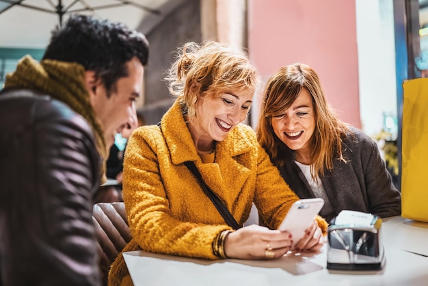 Group of cheerful adults sitting at cafe watching content on smartphone screen - Adult friend smiling browsing social network sitting at cafe outdoors