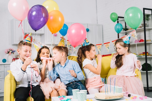 Group of children sitting on sofa holding colorful balloons and blowing party horn