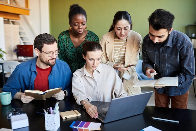 Group of creative designers gathered by table in front of laptop making notes while watching online