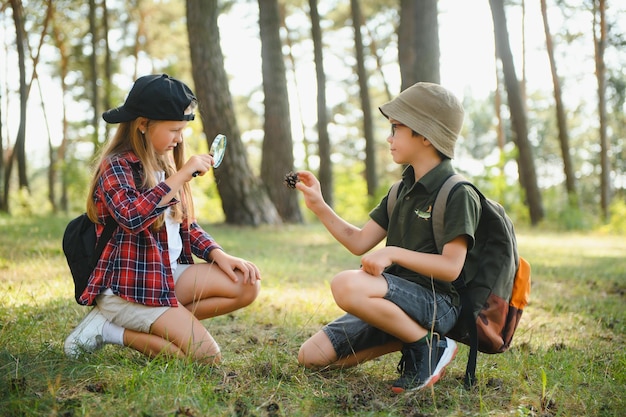 Group of curious happy school kids in casual clothes with backpacks exploring nature and forest together on sunny autumn day girl holding magnifying glass and looking at fir cone in boy hands