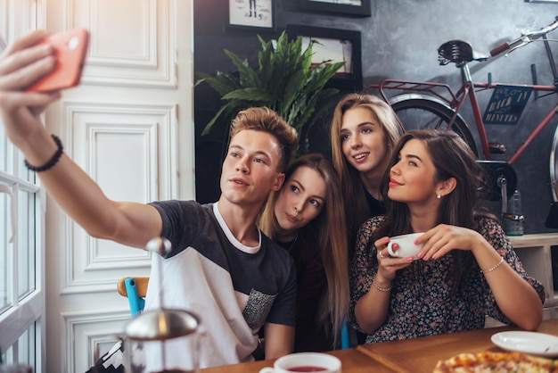 Group of cute teenagers taking selfie with cellphone while sitting in a restaurant with interior in retro style