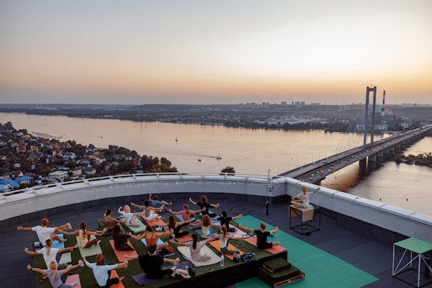 Group of different people are doing meditative yoga practices at the rooftop on a beautiful sunset at summer evening