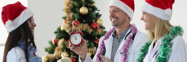 Group of doctors in santa claus hats standing near christmas tree and holding red alarm clock