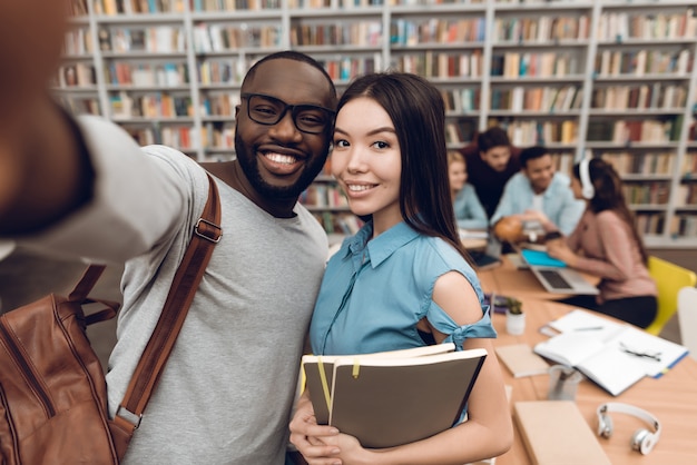 Group of ethnic multicultural students in library. Black guy and asian girl taking selfie on phone.