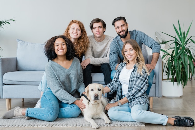 Group of five affectionate teenage guys and girls and white labrador sitting on the floor and on couch