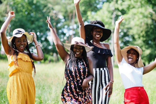 Group of four gorgeous african american womans wear summer hat spending time at green grass in park.