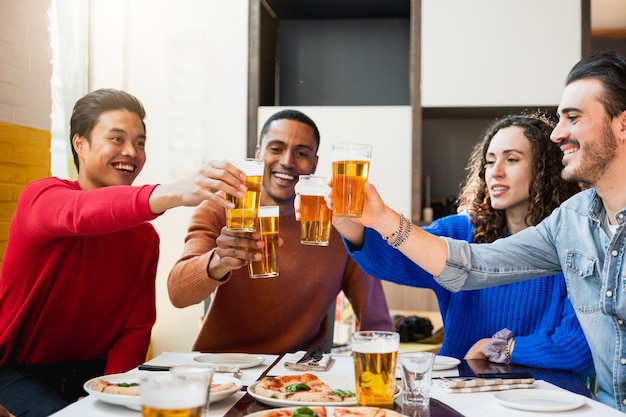 Group of four multiethnic friends eating pizza in a restaurant and cheering with glasses of beer