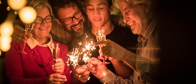 Group of four people together at night celebrating with sparlers and glass with wine or champagne - family of teenager, adult, and two seniors celebrating the new year