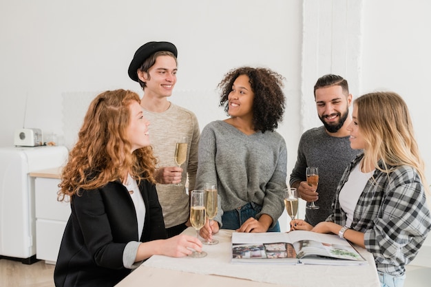 Group of friendly guys and girls with flutes of champagne chatting and looking through magazine at home party