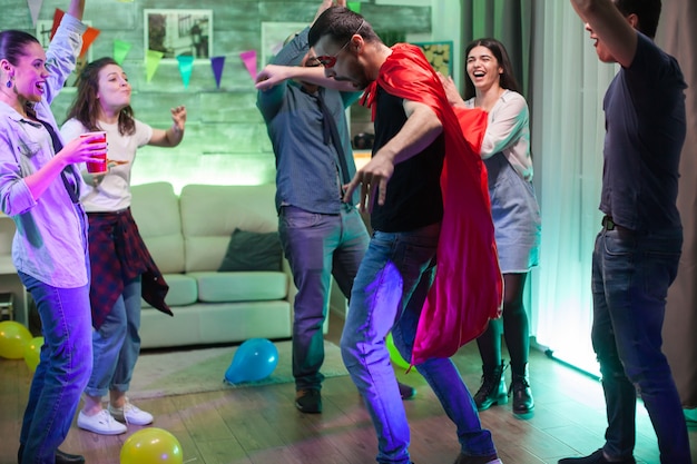 Group of friends cheering for their friend dressed up like a super hero at the party with red cape.